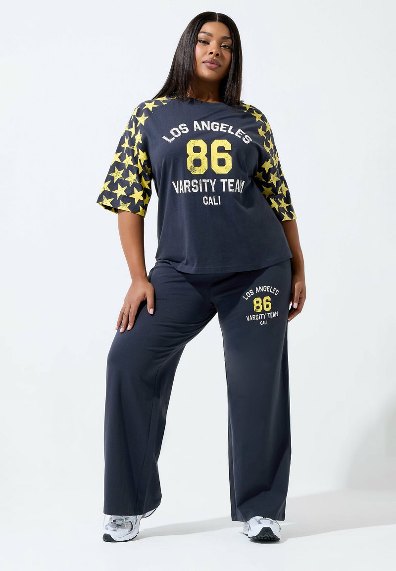 Frau in schwarzem Los Angeles-College-Outfit mit gelben Sternen auf den Ärmeln und weißen Turnschuhen, steht mit einer Hand in der Hüfte.