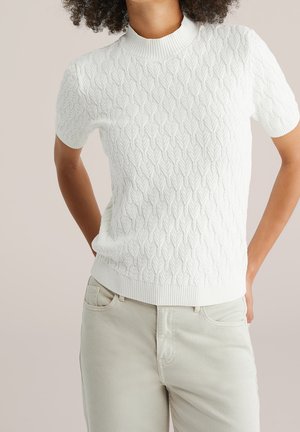 Valkoinen neulottu lyhythihainen villapaita, jossa on korkea ribbineck sekä teksturoitu lehtimäinen kuvio. Yhdistetty vaaleanvärisiin housuihin.