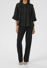 Blouse en coton noir avec manches évasées et plis à l'avant, portée avec un pantalon noir et des sandales. Col simple et fermeture à boutons.