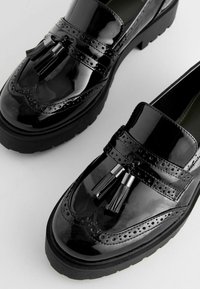 Sorte patenterede læderslippers med brogue-detaljer, der har en tyk sål og dekorative kvaster foran. Flad design og glat tekstur.