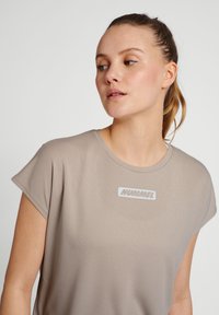 Beige t-shirt med kort ärm och mjuk textur, med en vit logotypapplikation som läser "HUMMEL" i ett rektangulärt mönster.