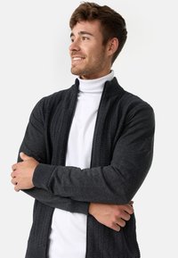 Mørkegrå zip-up sweater prydet med et struktureret strikdesign foran, glatte ærmer og ribbede manchetter. Bæres over en hvid turtleneck.