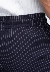 Gros plan de la taille portant un pantalon bleu marine à fines rayures verticales blanches et un top en maille côtelée blanche.