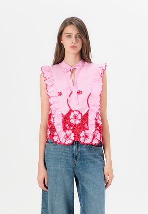 Ermeløs rosa bluse med volangkrage, blomsterlacet design i rødt, og knyting rundt halsen. Kombinert med løse blå denimjeans.