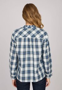 Chemise boutonnée à carreaux bleus et blancs avec manches longues, col plat, et accents réfléchissants sur les épaules et les poignets, fabriquée en tissu tissé.