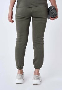Pantalones de chándal tapered en color verde oliva con puños elásticos en los tobillos, combinados con zapatillas deportivas blancas. La tela parece suave y flexible.