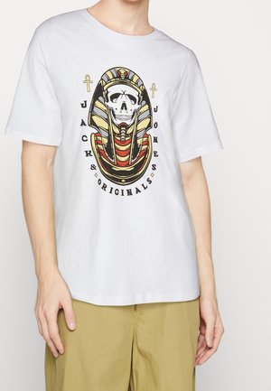 Osoba ubrana w białą koszulkę z grafiką czaszki faraona, dwoma symbolami ankh i napisem „"JACK & JONES ORIGINALS"”, zestawioną z jasnobrązowymi spodniami.