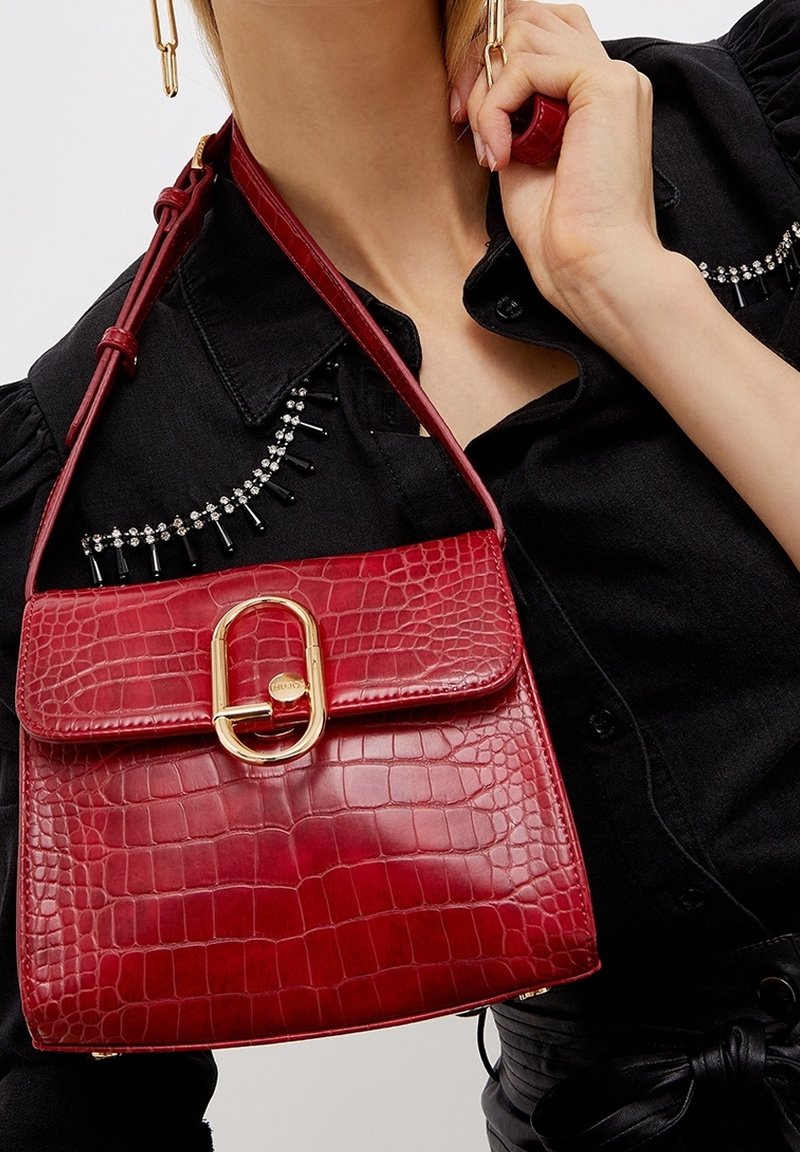 Femme tenant un sac à main en cuir rouge texturé crocodile avec boucle ovale dorée, portant une chemise noire avec des détails en strass et perles noires.