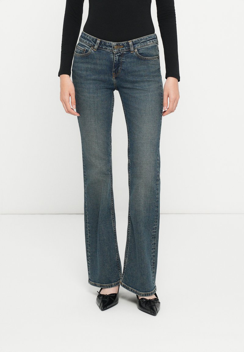 Bukser i blå denim med vidde, klassisk passform, fem lommer og metallknaplukning. Stoffet har en jevn tekstur med subtil falming.