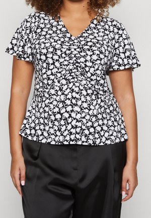 Mujer con una camiseta de manga corta floral en blanco y negro con fruncido en el delantero, combinada con pantalones de satén negros.