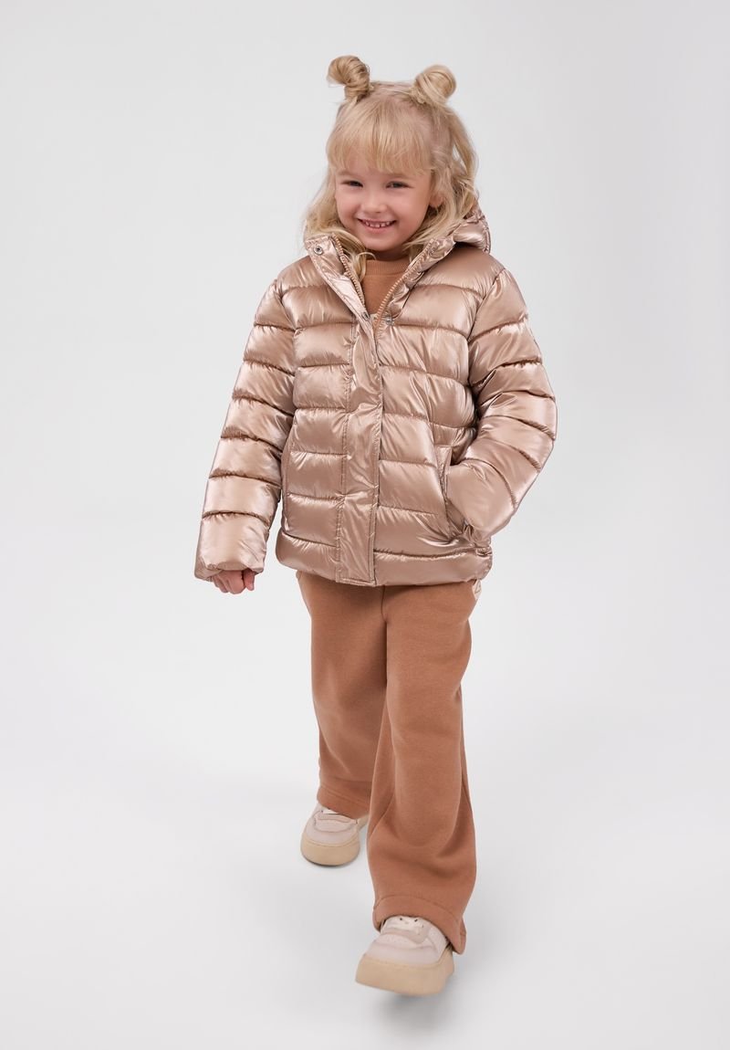 Jeune fille souriante aux cheveux blonds coiffés en deux chignons porte une doudoune beige brillante, un pantalon marron et des baskets blanches en marchant vers l'avant.