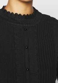Gros plan sur un cardigan noir tricoté avec un col festonné et trois boutons noirs sur le devant.