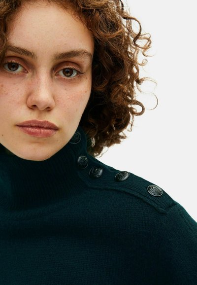 Mørkegrøn højhalset sweater med strikket tekstur, der har sorte dekorative knapper på venstre skulder og en ribbenkrave.