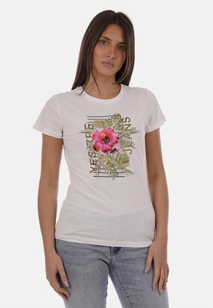 Giovane donna che indossa una maglietta bianca con una grande stampa di un fiore rosa e foglie verdi, abbinata a jeans azzurri, in piedi contro uno sfondo bianco.