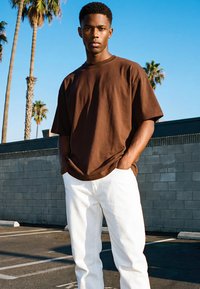 Fiatal férfi barna pólóban és fehér nadrágban áll, kezét a zsebében tartva egy napos, szabadtéri parkolóban, pálmafák és kék ég alatt.