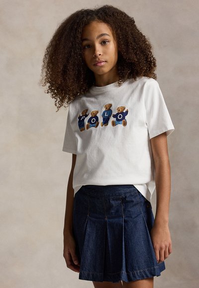 Νεαρή κοπέλα με σγουρά μαλλιά που φοράει ένα λευκό μπλουζάκι με τέσσερις αρκούδες που σχηματίζουν τη λέξη «POLO» και μια σκούρα μπλε πλισέ τζιν φούστα.