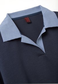 Camisa azul marino con un cuello doblado que presenta un patrón de cuadros pequeños en azul y blanco, y una etiqueta de marca roja y negra en el interior del cuello.