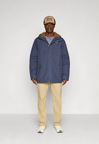 Marineblå isolert jakke med pelsforet hette, to frontlommer og et grått innerlag, kombinert med kremfargede bukser og hvite joggesko.