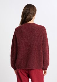Maglione lavorato a maglia bordeaux con una finitura texturizzata e pelosa, caratterizzato da un collo a giro e spalle scese. Dettagli cuciti lungo le cuciture.