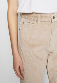 Zblízka je vidět osobu ve béžových manšestrových kalhotách a bílé košili, s uvolněnou levou rukou a rukou podél těla.