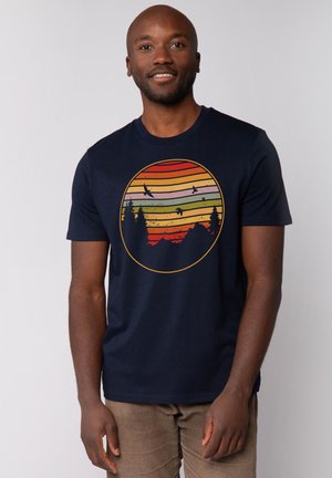 Marineblauwe t-shirt met korte mouwen, voorzien van een cirkelvormige afbeelding van een zonsondergang met horizontale strepen in oranje, geel en groen, en silhouetten van bomen.