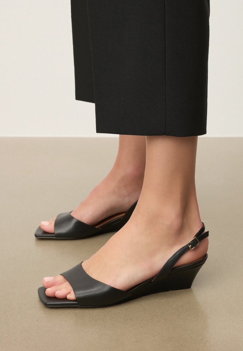 Picioare purtând sandale negre cu platformă și bretele din piele, cu degetele pătrate, asortate cu pantaloni negri scurți, pe un podea bej.