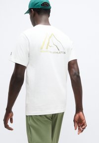 Λευκό βαμβακερό t-shirt με κοντά μανίκια που διαθέτει χρυσό γραφικό βουνού και τη φράση «MOUNTAIN ATHLETICS» στην πλάτη. Φορά πράσινο καπέλο.