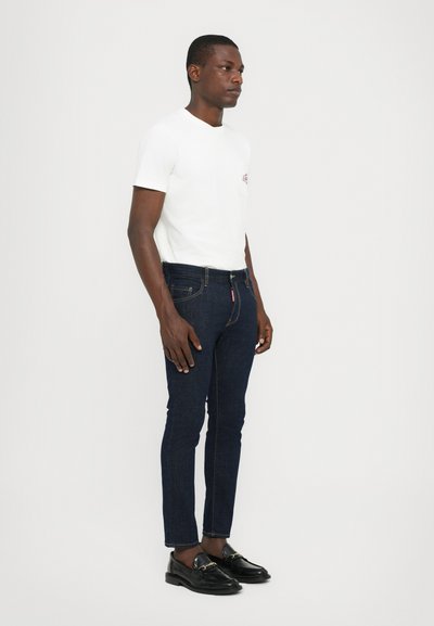 Maglietta bianca a maniche corte con una piccola grafica, abbinata a jeans slim-fit blu scuro e mocassini neri con accenti dorati.