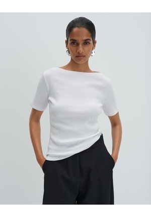 Femme portant un haut blanc à manches courtes et un pantalon noir, debout les mains dans les poches devant un fond gris uni.