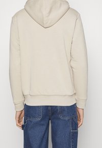 Sweat à capuche beige avec une poche kangourou, des poignets et un ourlet côtelés. Le tissu semble doux et texturé, conçu pour le confort.