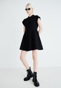Rochie neagră, cu butoane, mâneci scurte rulate, dintr-un material texturat și fustă evazată. Asortată cu cizme negre cu șireturi până la gleznă.