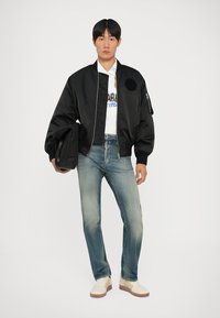 Giacca bomber nera con una toppa rotonda, camicia bianca con logo, jeans azzurri chiari e sneakers beige. Modello che tiene una borsa nera.