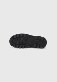 Semelle de chaussure en caoutchouc noir avec une surface texturée et des rainures profondes, comportant divers motifs géométriques pour l'adhérence et la traction.