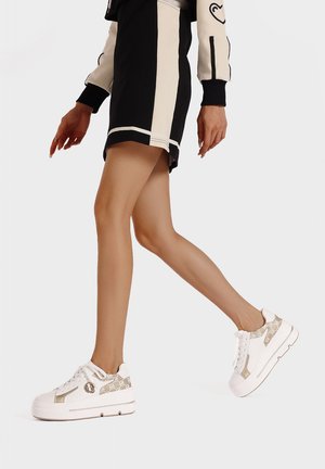 Sneakers bianchi con un pannello laterale beige testurizzato, punta rotonda, suola piatta e design con lacci, posizionate su uno sfondo minimalista.