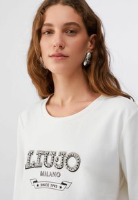 Jeune femme aux longs cheveux bruns portant un sweat-shirt blanc avec le logo orné "LIU JO Milano Since 1995" et des boucles d'oreilles argentées en anneau.