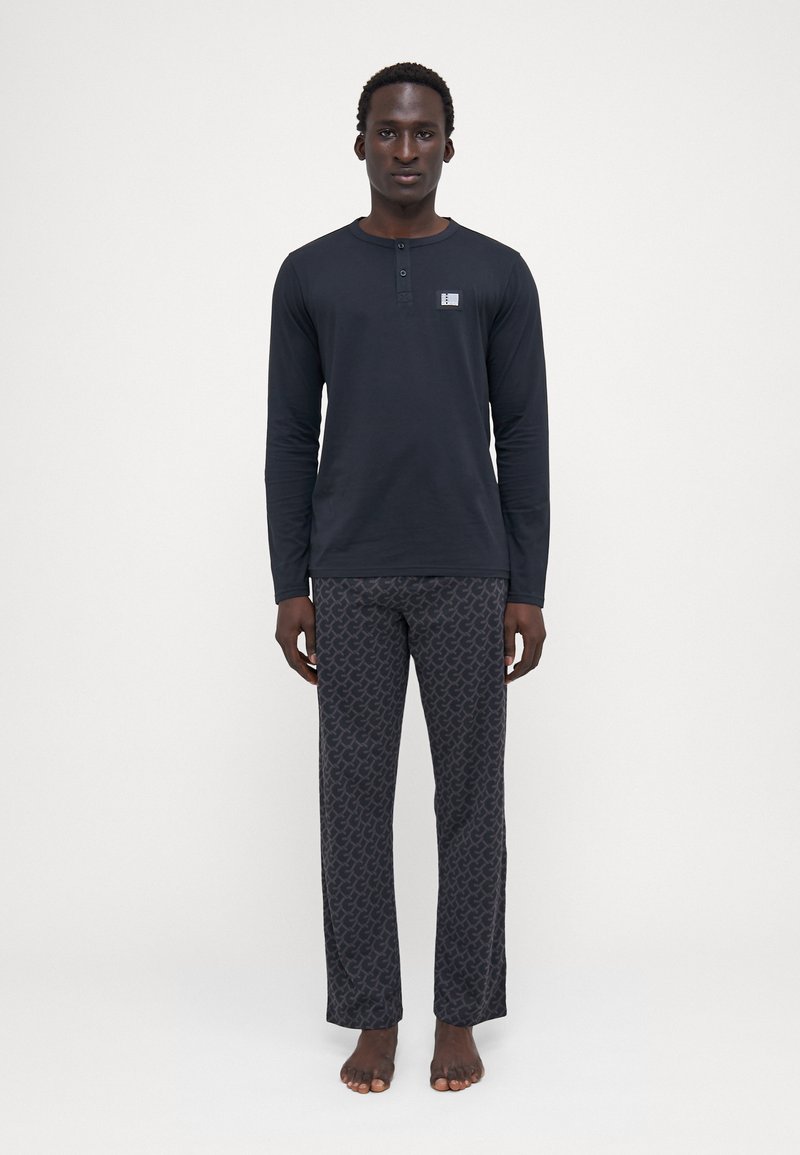 Camisa henley negra de manga larga con detalles de botones, combinada con pantalones de salón grises con un patrón sutil repetitivo.