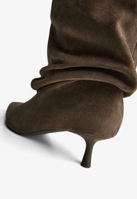 Stivaletto alla caviglia con tacco in pelle scamosciata marrone, dalla punta appuntita, caratterizzato da un design superiore morbido e pieghettato e un tacco sottile e affusolato. Texture morbida in tutto.