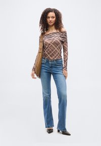 Γυναίκα που στέκεται, φοράει μπλούζα με καρό σχέδιο και έξω από τον ώμο, μπλε τζιν, μαύρα μυτερά παπούτσια και καφέ τσάντα ώμου σε λευκό φόντο.