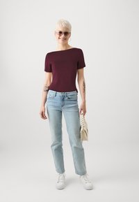Maronnsfarget kortermet topp, lyseblå jeans, hvite joggesko, holder en beige veske. Tatoveringer synlige på armene, har på seg runde solbriller.