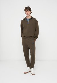 Oljčno zelena trenirka z enojnim zadrgo in modro oznako, združena z usklajenimi trenirkami. Bele superge rjave podplate. Enostaven dizajn.