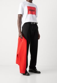 Camiseta blanca con gráfico rectangular rojo que dice "HUGO", pantalones negros, cinturón negro, sosteniendo un suéter rojo, zapatos negros, texturas suaves.