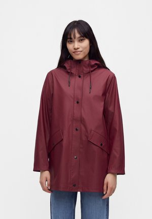 Femme portant un imperméable à capuche bordeaux avec des boutons à l'avant et des poches à rabat, debout devant un fond uni.