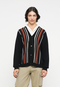 Zwarte cardigan met meerkleurige verticale strepen in rood, wit en beige. Kenmerkt zich door een v-hals, knopen en geribbelde manchetten en zoom.
