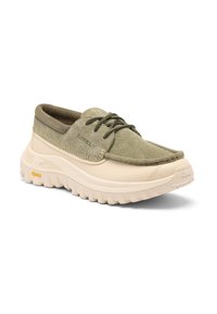 Chaussure beige et vert olive en suède à lacets, avec une semelle blanche épaisse et texturée et un logo Vibram sur le côté, inclinée pour montrer le côté extérieur et l'avant.