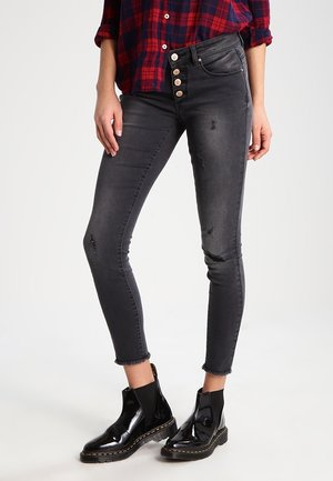 Vrouw draagt zwarte versleten skinny jeans met zichtbare knopen, rafelige zomen, zwarte glanzende enkellaarzen en een rood geruit overhemd.