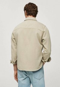 Camisa de color beige con puños abotonados y cuello de punta. Jeans de denim claros visibles a la altura de la cintura, con bolsillos traseros visibles.