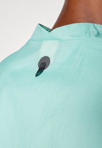 Light aqua shirt with a textured grid pattern. It features a back neck closure with a round black button and a small black loop.