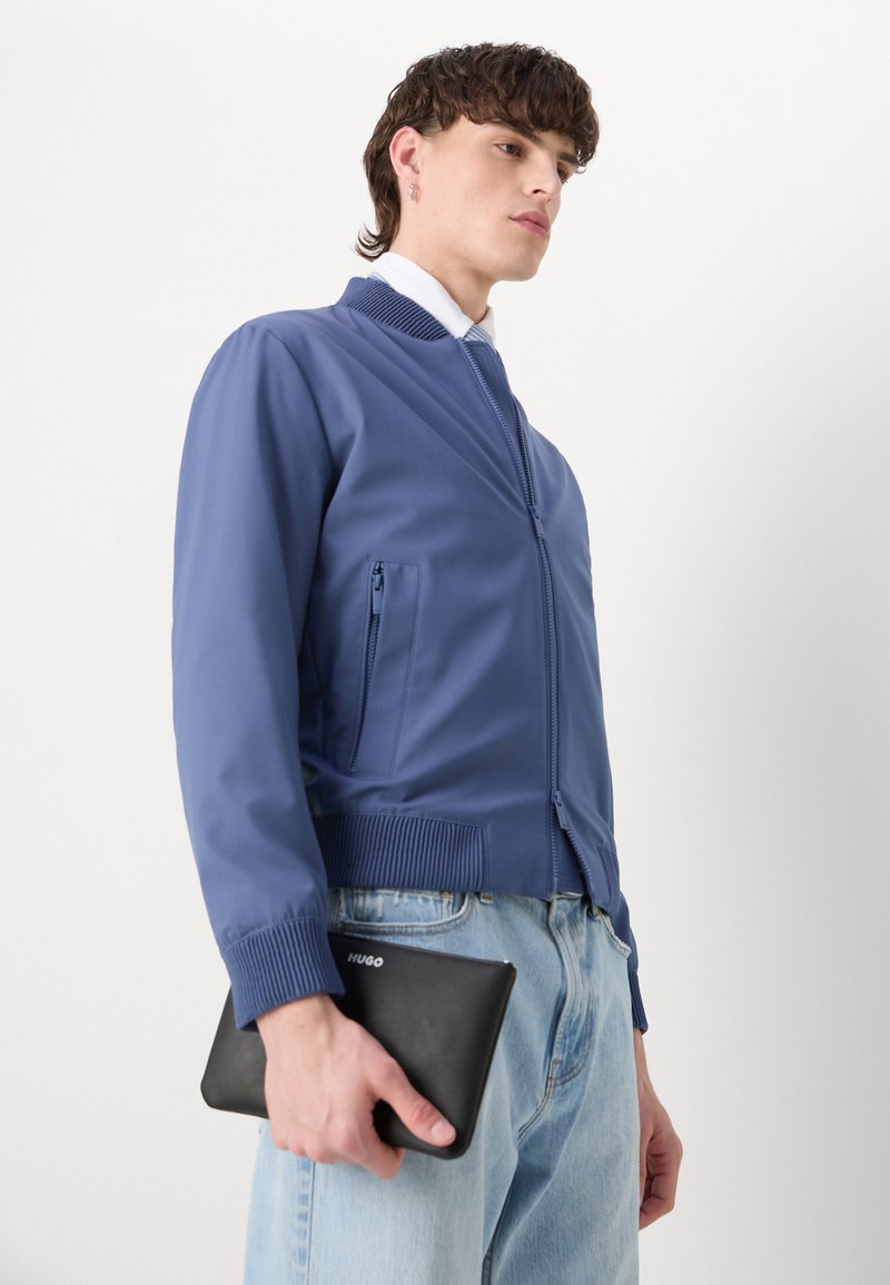 Veste bomber bleue avec des poignets et un col côtelés, poche zippée, portée avec un jean en denim clair. Tenant une pochette en cuir noir.
