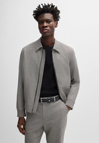 Grå mönstrad zip-up jacka med krage, kombinerad med en svart texturerad skjorta och grå byxor. Har elastiska manschetter och ett bälte.