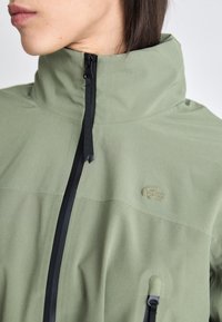 Giacca verde con colletto alto, zip nera e logo tono su tono. Presenta una texture liscia e una tasca frontale funzionale.
